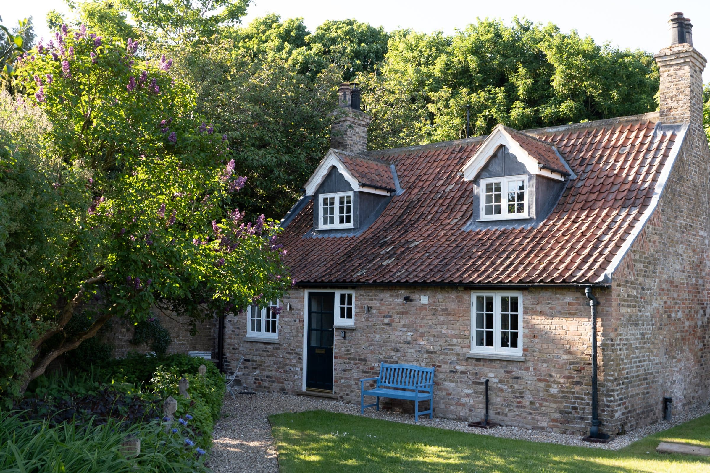 The Cottage with its pantiled roof and dormers, a blue bench by the dark front door and lilac in flower.