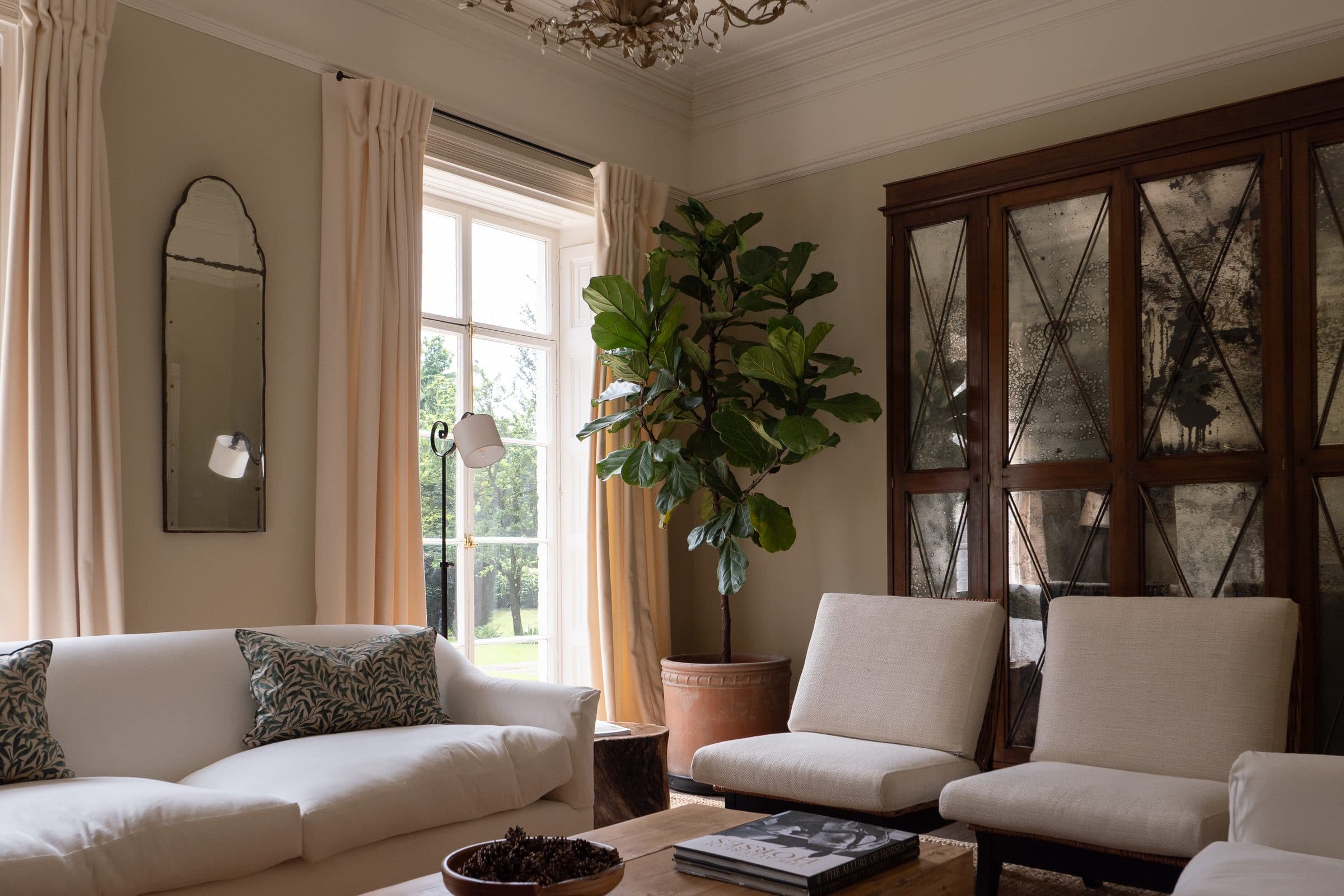 The drawing room with a cream sofa, fiddle-leaf fig and antiqued-mirror cabinet beside a tall sash window onto the garden.