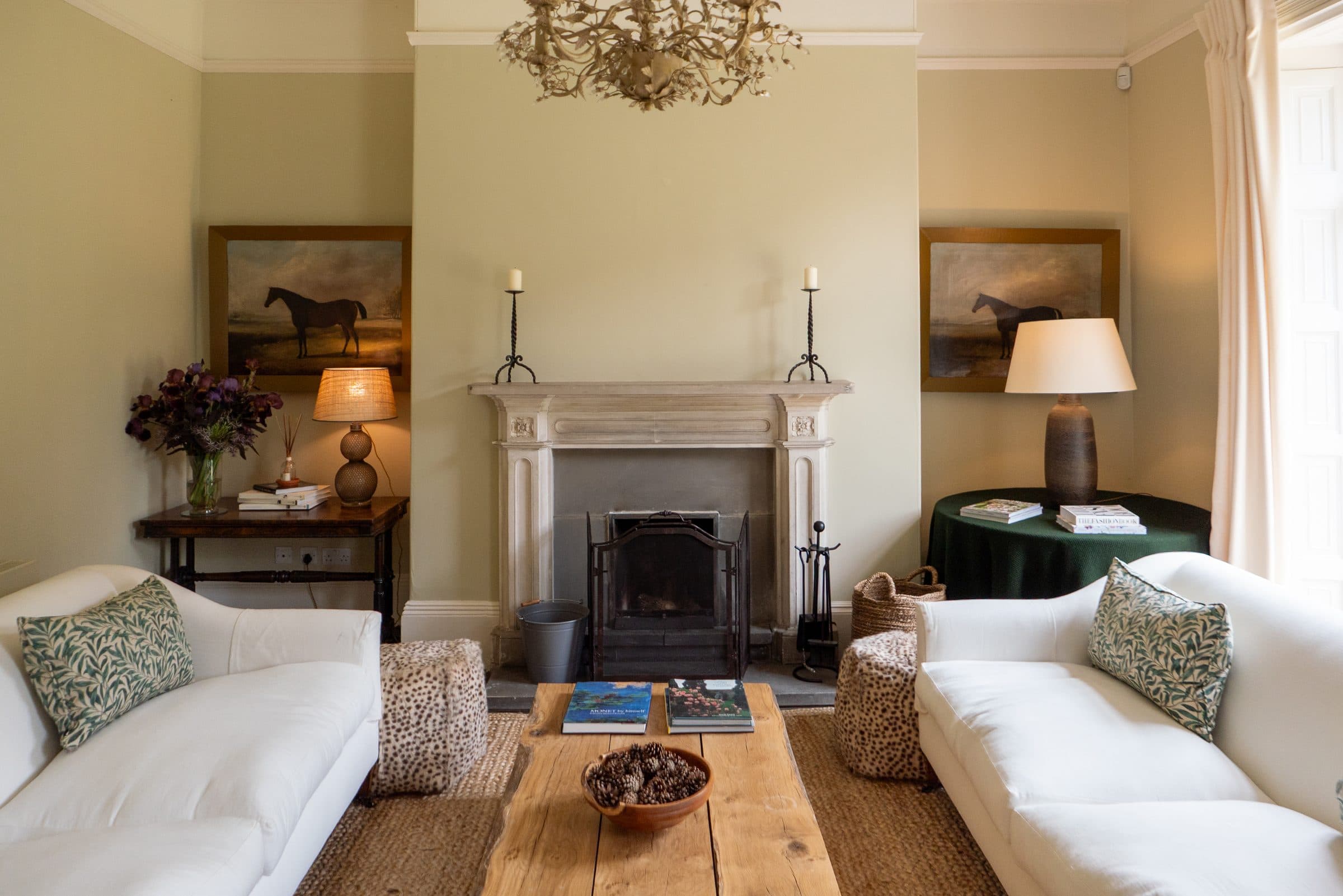 Two white sofas facing across a rustic oak table towards the stone fireplace, hung with an antique painting of a horse.