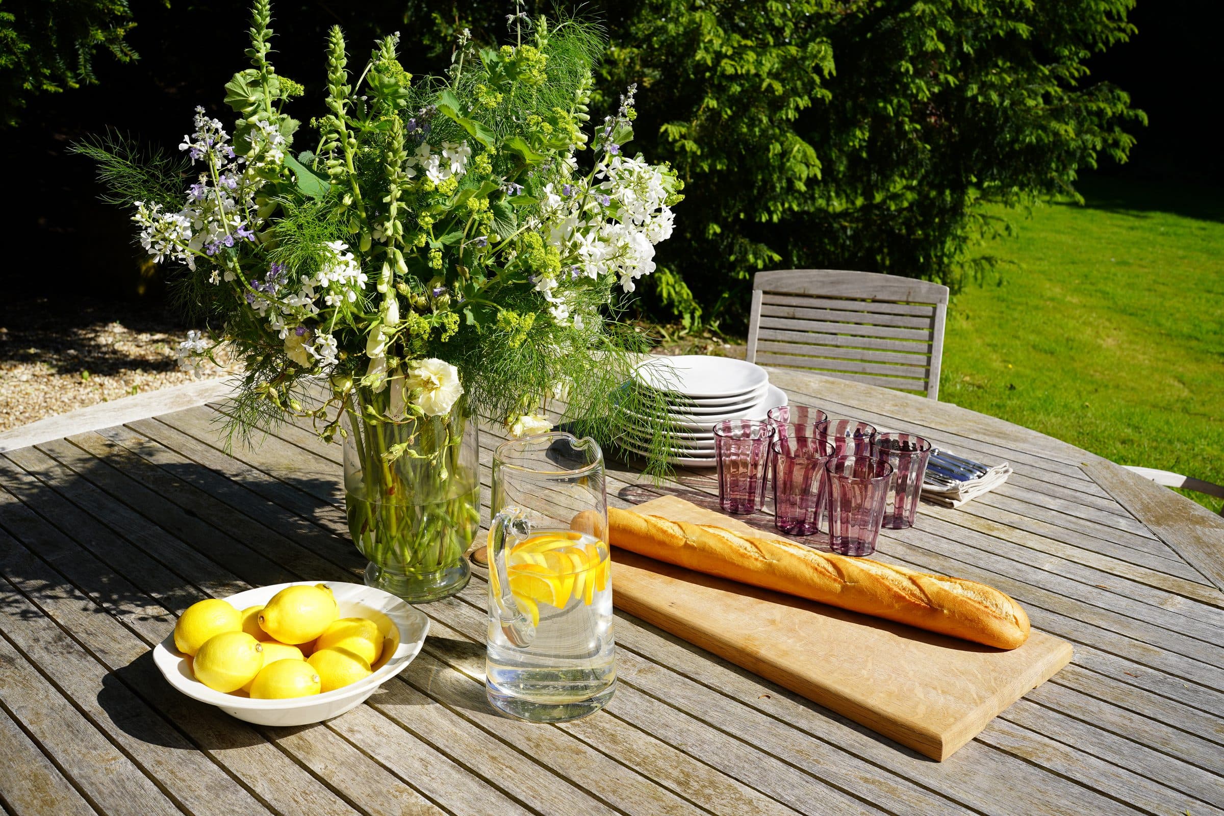 A weathered teak garden table set with lemons, green glassware, a baguette and a jug of wild flowers on the lawn.