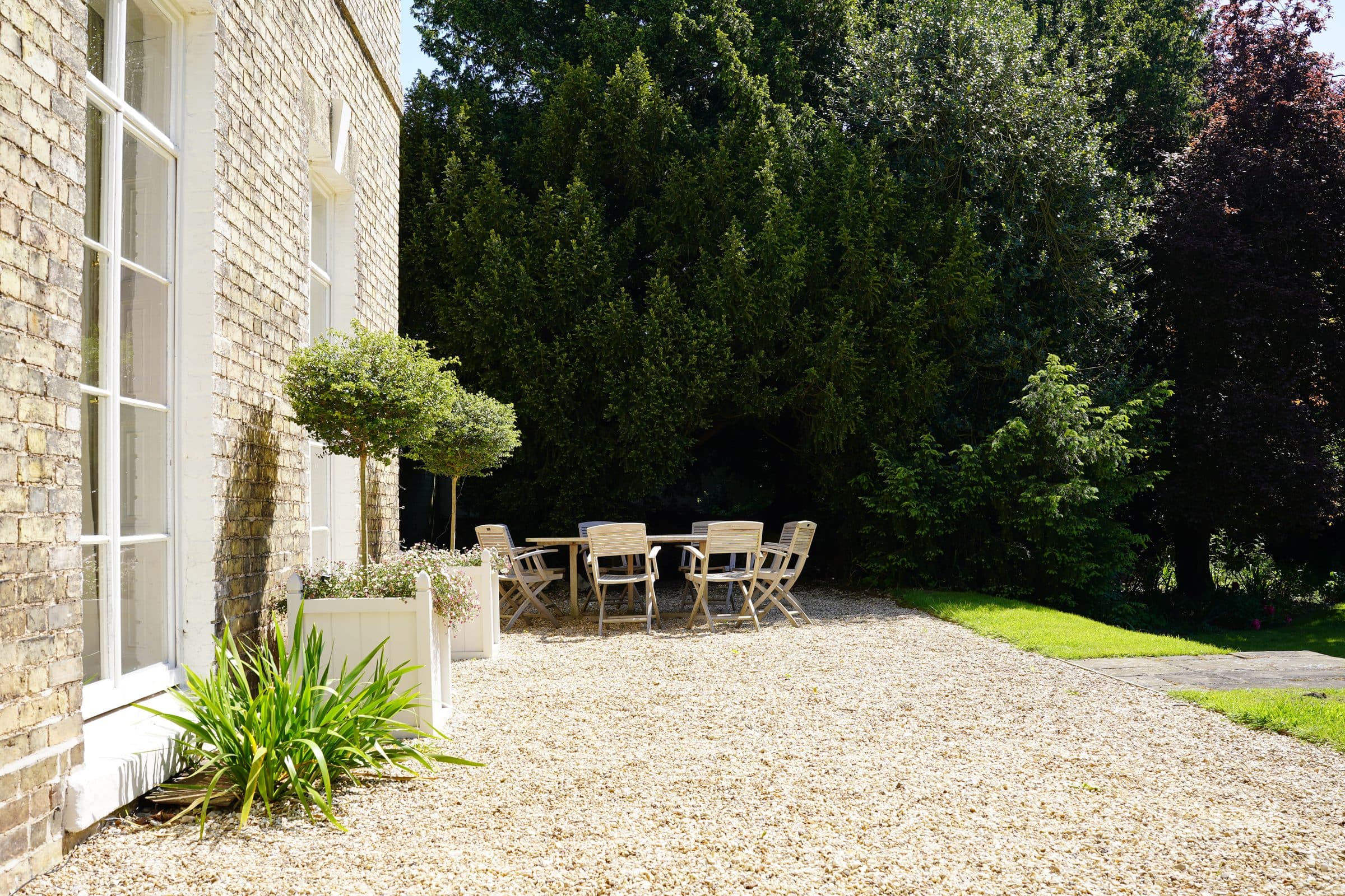 The gravel terrace beside the house, with an outdoor table set under the trees.