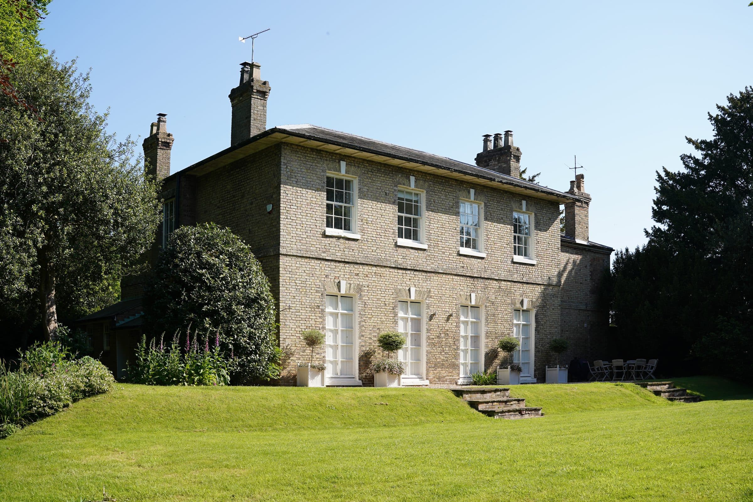 The vicarage's brick garden elevation with tall sash windows, seen across the sunlit lawn on a clear summer morning.
